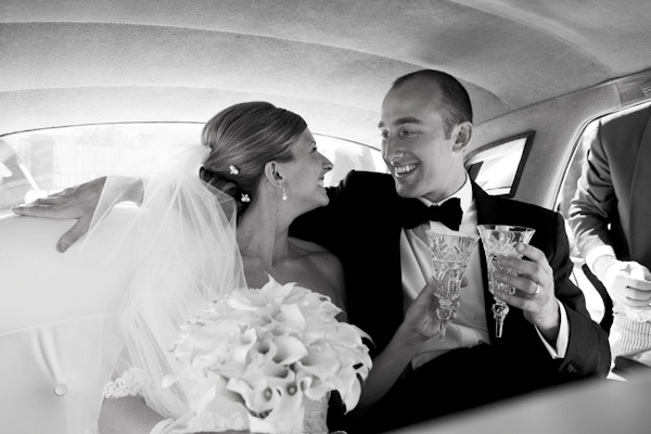 black and white photo of bride and groom toasting in the back of limousine as they get ready to leave the ceremony -  bride is wearing full length veil with hair in a low bun and groom is wearing a tuxedo - wedding photo by Jennifer Bowen Photography